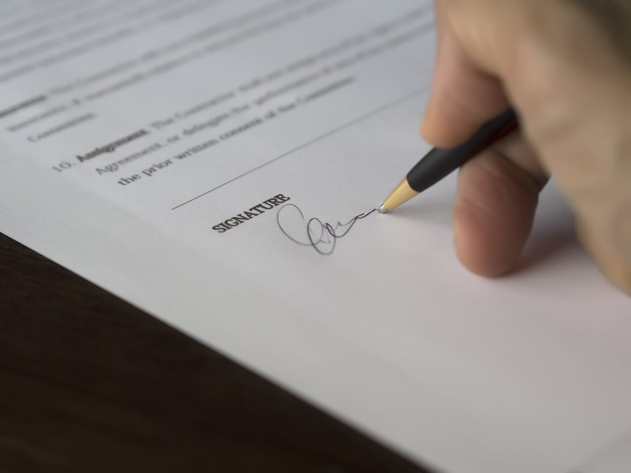 about-img A hand signs a formal contract with a pen on a wooden desk.